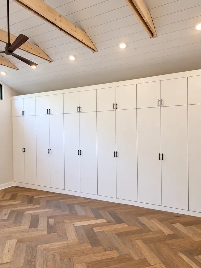 Wall of white flat-panel built-in wardrobes with black pulls under a beamed ceiling, above a herringbone wood floor