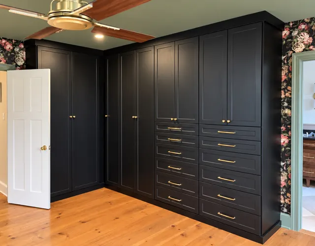 Black L-shaped built-in wardrobes with shaker doors, stacked drawers and brass pulls in a floral-papered bedroom