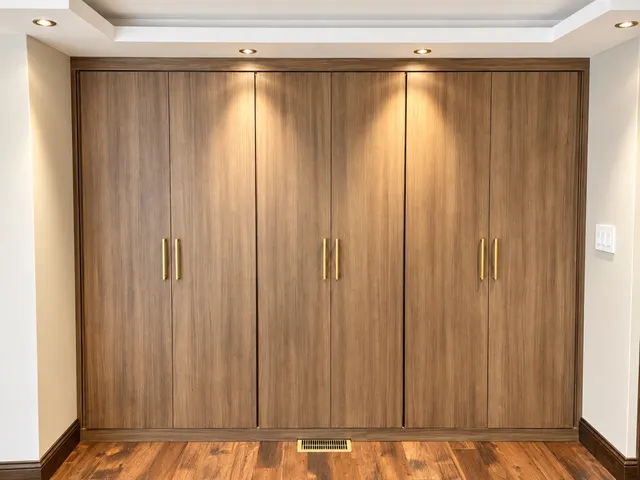Walnut-finish built-in wardrobe with six tall flat-panel doors and brass pulls under recessed lights