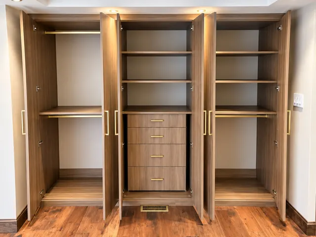 Walnut-finish built-in wardrobe with doors open to lit shelves, brass hanging rods and a four-drawer center stack