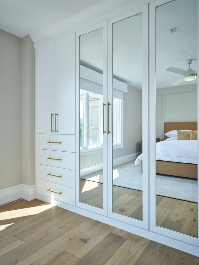 White built-in wardrobe with an upper cabinet, four drawers and full-length mirrored doors with brass pulls