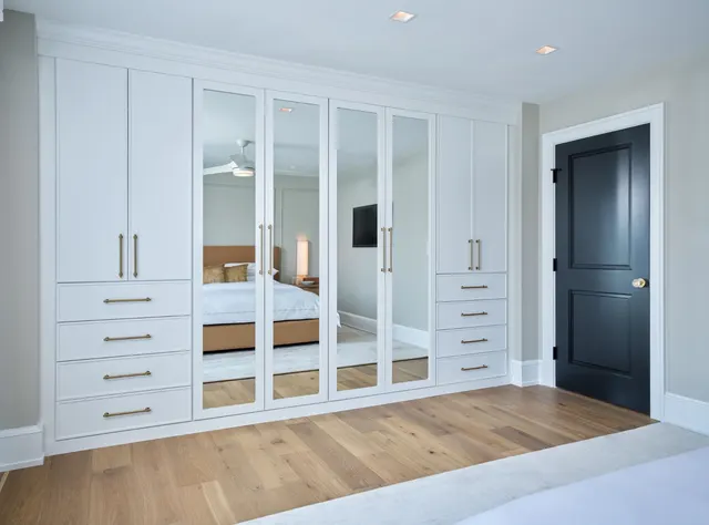 White bedroom wardrobe wall with four mirrored center doors between cabinets and drawer stacks with brass pulls