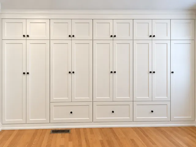 Wall of white shaker built-in wardrobes with black knobs, upper cabinets and three wide drawers