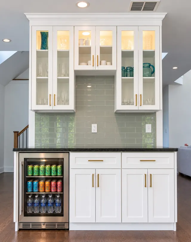 White shaker beverage station with lit glass-front cabinets, green glass tile, a dark counter and a beverage fridge