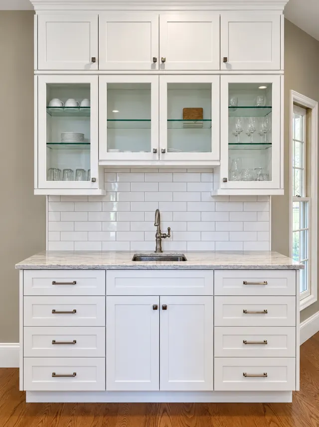 White shaker beverage station with glass-front upper cabinets, subway tile, a marble counter and a small sink