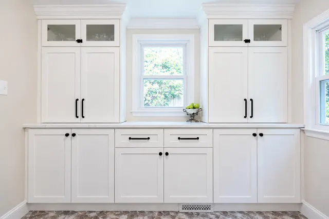 White shaker buffet with glass-front upper cabinets on each side of a window, a light counter and black hardware