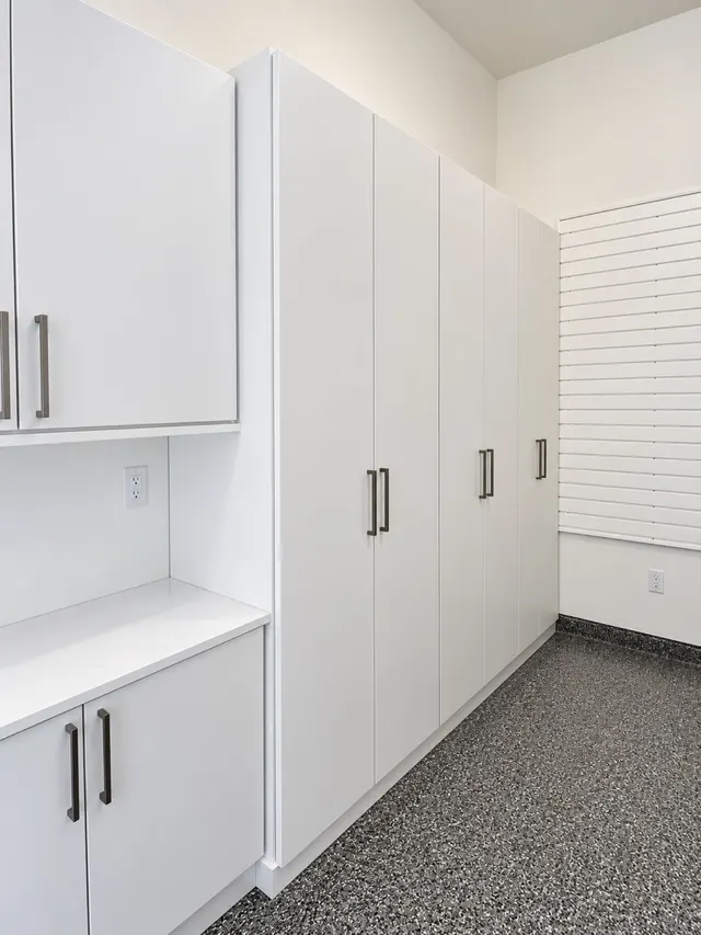Tall white garage cabinets with bronze pulls beside a counter and white slatwall, on a speckled epoxy floor
