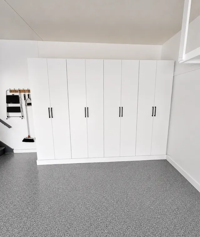 Row of tall white garage cabinets with black pulls on a speckled epoxy floor, with wall hooks holding a step stool and broom