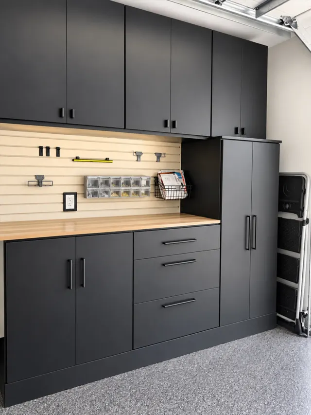 Black garage cabinets and drawers with a butcher block workbench and slatwall holding hooks, bins and a basket