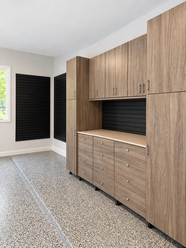 Wood-grain garage cabinets and drawer banks with a light counter and black slatwall, with more slatwall near a window