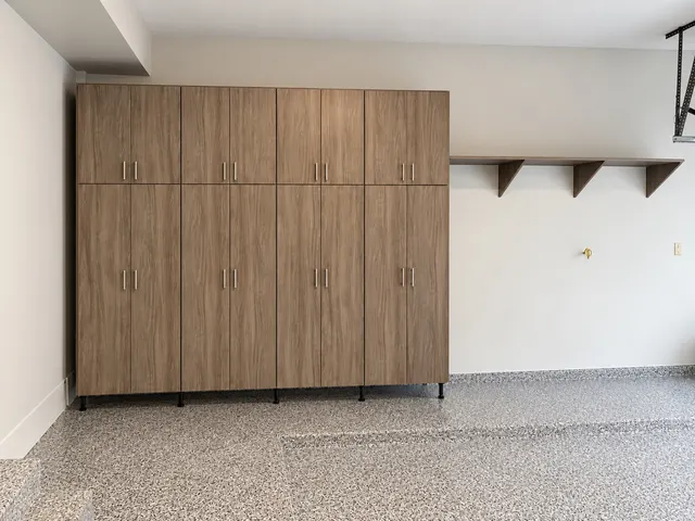 Wall of tall wood-grain garage cabinets on legs beside a long bracketed wall shelf, on a speckled epoxy floor
