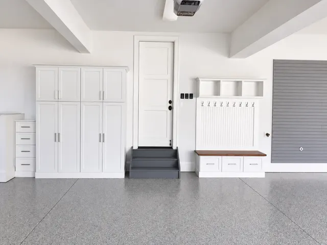 White garage cabinets and a bench with hooks and cubbies beside a door with gray steps, next to gray slatwall