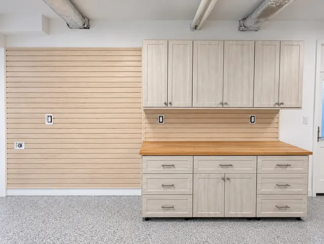 Light wood-grain garage cabinets and drawers with a butcher block top beside a wood-look slatwall wall
