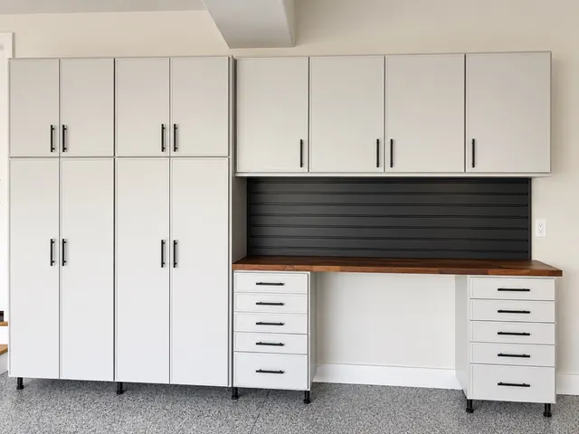 White garage cabinets with black pulls over a wood-top workbench, drawer stacks and black slatwall backsplash