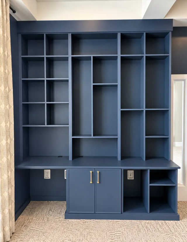 Navy built-in office wall with varied open shelving, a desk surface, cabinet doors with brass pulls and open cubbies