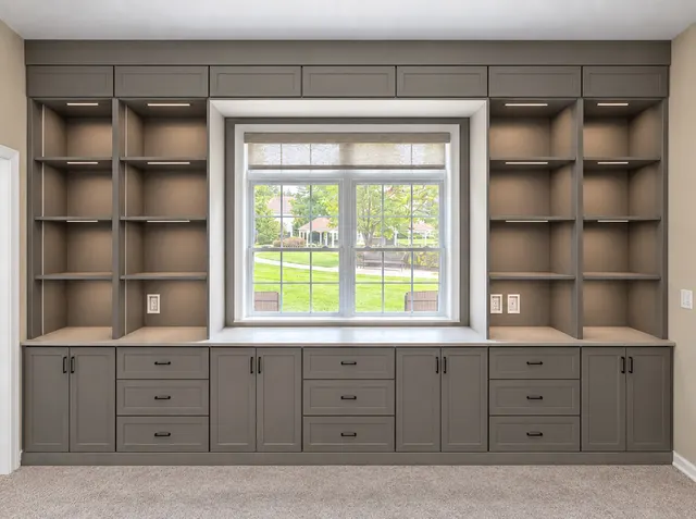 Gray built-in bookcases with lit shelves and upper cabinets surrounding a window, over a row of cabinets and drawers