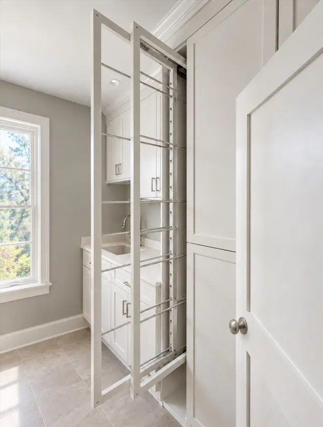 Tall pull-out drying rack with metal rods extended from white laundry cabinets, beside a sink counter and window