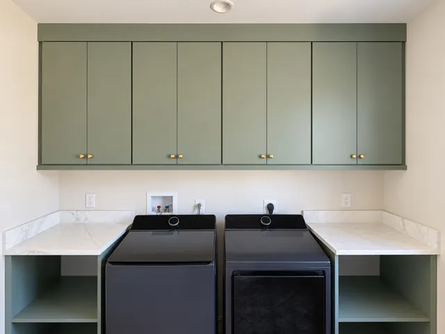Green flat-panel wall cabinets with brass knobs above a top-load washer and dryer, flanked by marble-look counters over open green shelves