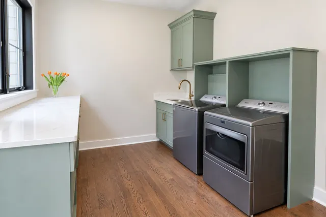 Sage green laundry room with a long white counter under a window, a sink with a brass faucet and an open green surround over a top-load washer and dryer