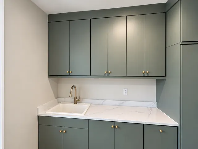 Green flat-panel laundry cabinets with brass knobs around a marble-look counter and a sink with a brass faucet