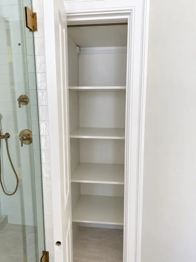 Narrow white linen closet with shelving beside a glass shower