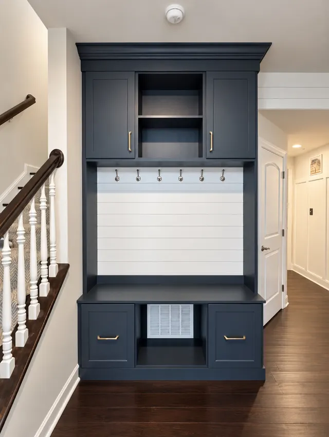 Navy entry locker beside a staircase with upper cabinets, a center cubby, a row of hooks on a white shiplap back, a bench and two drawers with brass pulls