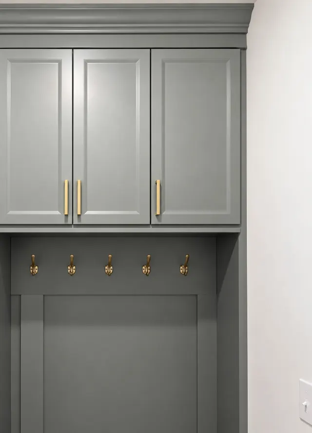 Close-up of gray-green upper cabinets with brass bar pulls above five brass hooks on a paneled back