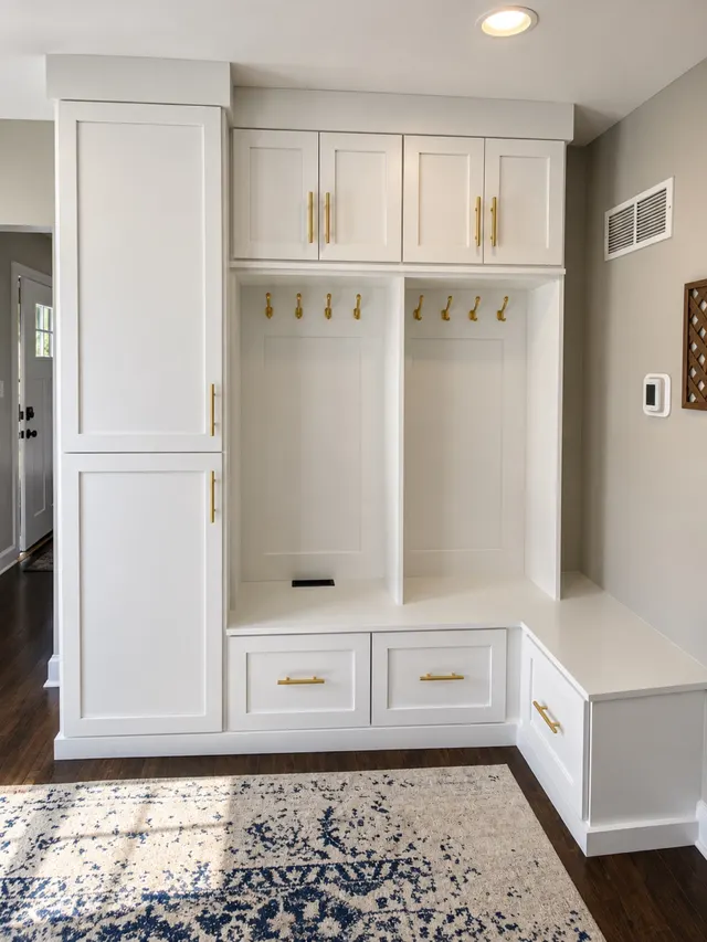 White L-shaped mudroom with a tall cabinet, two hook bays under upper cabinets and a corner bench over drawers with brass pulls