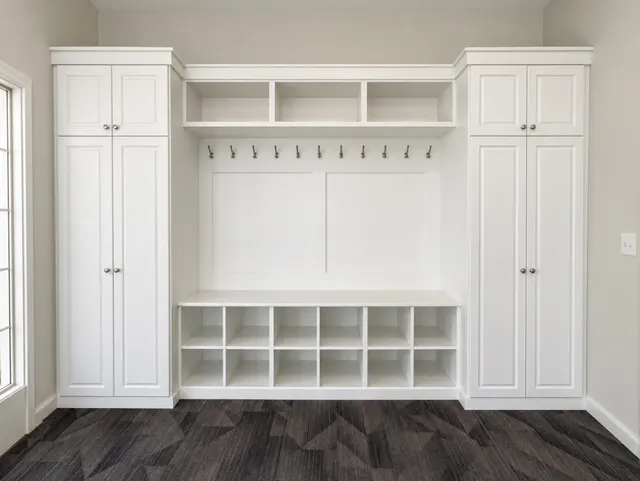 White mudroom wall with tall cabinets on each side of a row of hooks, open cubbies above and a bench over twelve shoe cubbies