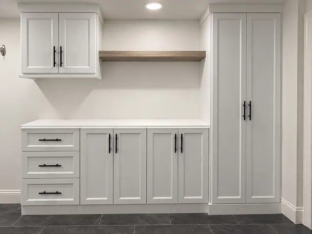 White mudroom cabinets and drawers with black bar pulls, a wood floating shelf and a tall two-door cabinet on a dark tile floor