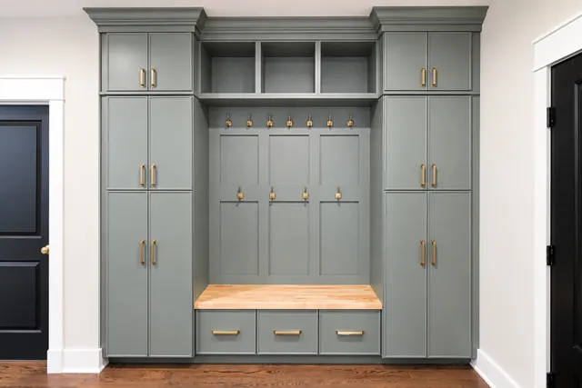 Green-gray mudroom lockers with brass hooks and pulls, a butcher-block bench over three drawers and tall cabinets on each side