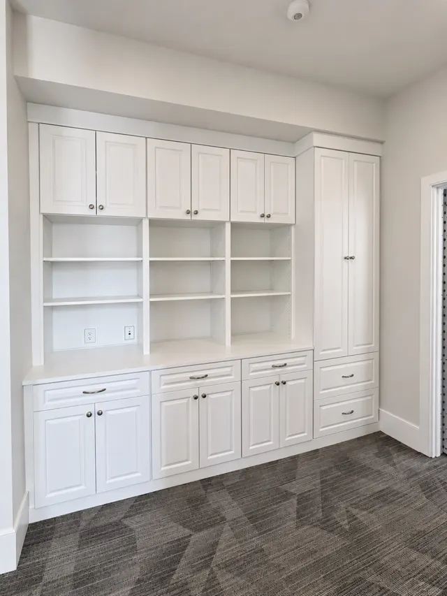 White wall of upper cabinets, open adjustable shelves and base cabinets with drawers beside a tall cabinet, on patterned carpet