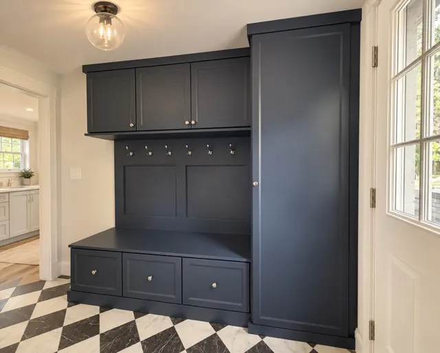 Navy mudroom unit with upper cabinets, six hooks over a paneled back, a bench over three drawers with nickel knobs and a tall cabinet, on a black-and-white checkered floor