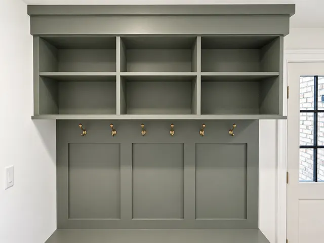 Sage green mudroom built-in with six open cubbies above a row of six brass hooks and a paneled back