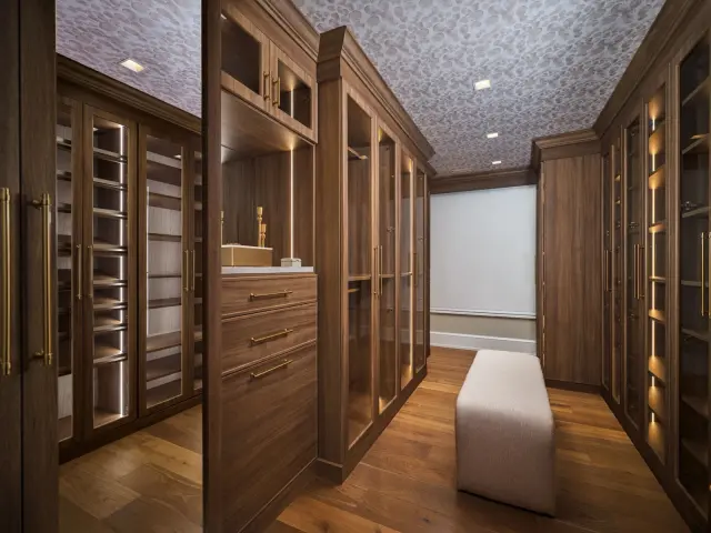 Dark walnut walk-in closet with lit glass-door cabinets and an upholstered ottoman