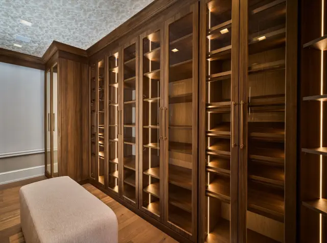 Walnut closet wall of illuminated glass-front cabinets with an ottoman