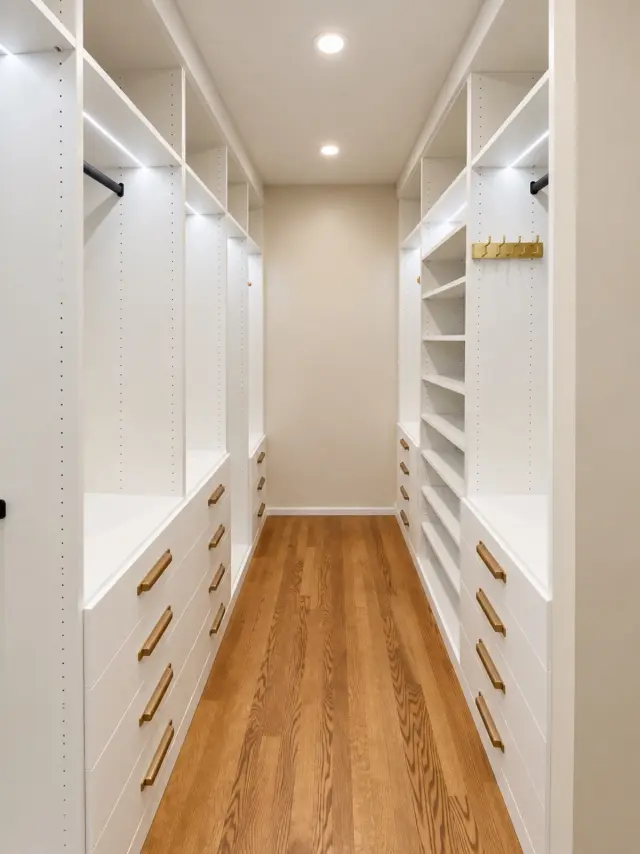 Narrow white walk-in closet with lit shelving, brass rods and drawers along both walls