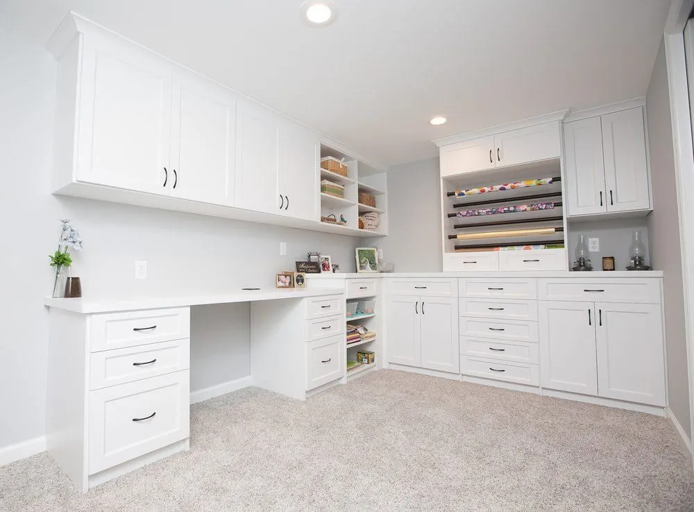 White L-shaped office with a desk, upper cabinets, drawers and a wrapping paper rack