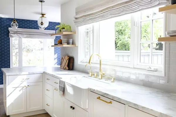 White kitchen sink under a window with brass fixtures and marble counters