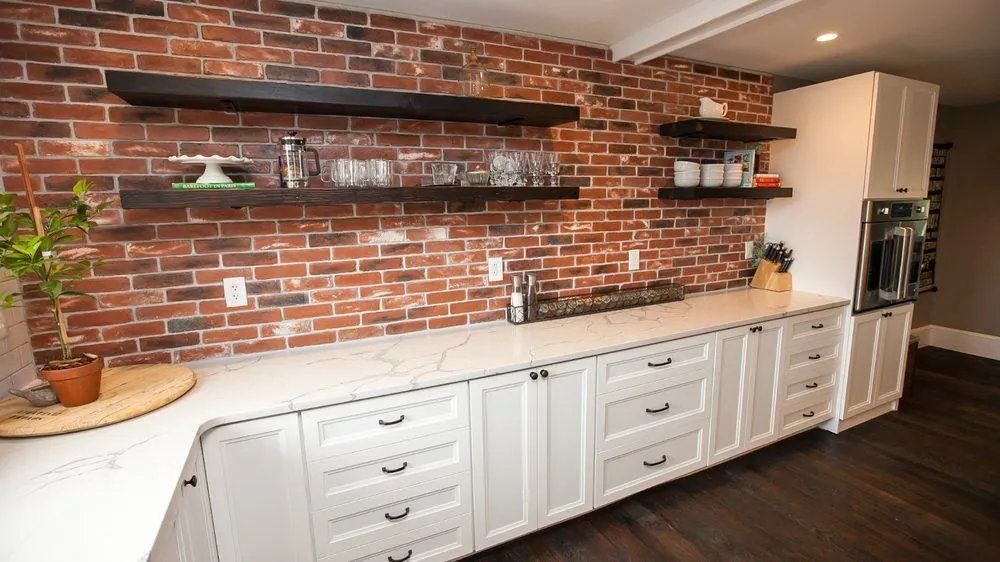White kitchen cabinets and a marble counter under open shelves on a brick wall