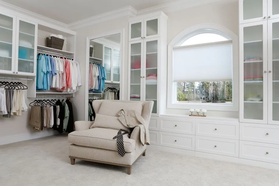 White closet with glass-door cabinets and a chaise under an arched window