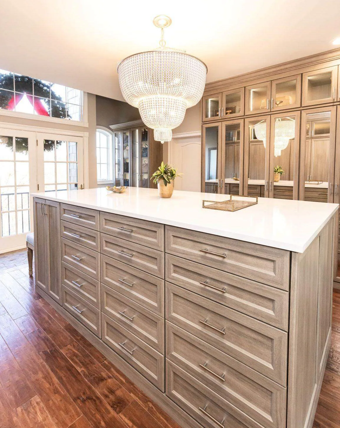 Walk-in closet island with drawers under a crystal chandelier, with glass-front cabinets