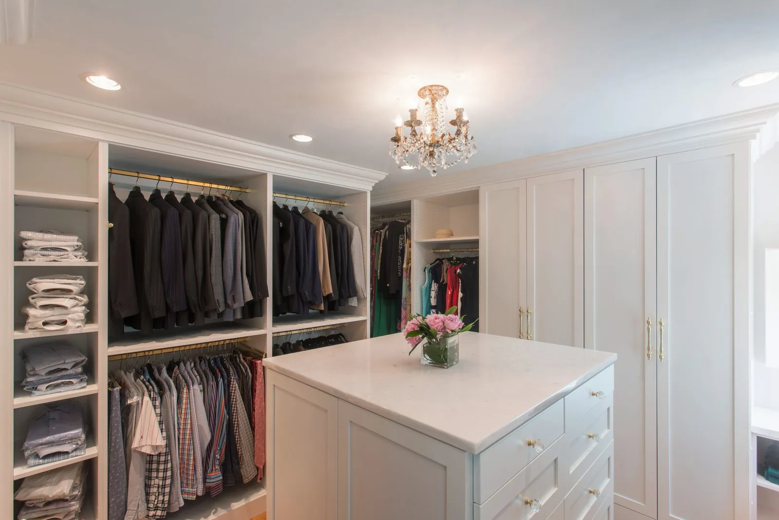 White walk-in closet with hanging, shelving and an island under a chandelier