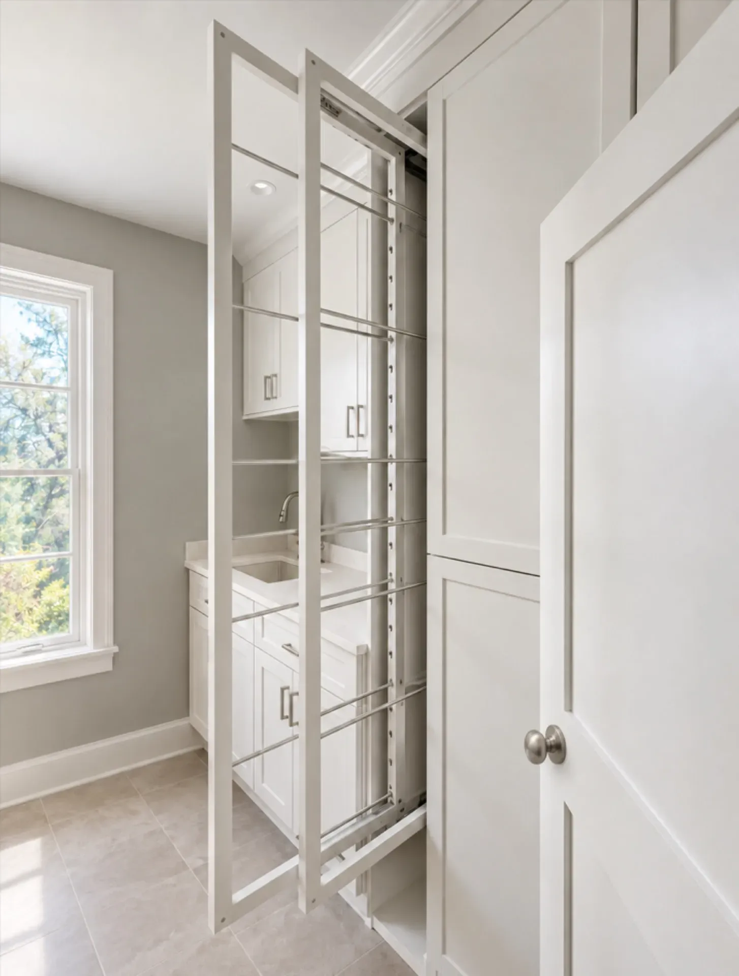 Tall pull-out drying rack with metal rods extended from white laundry cabinets, beside a sink counter and window